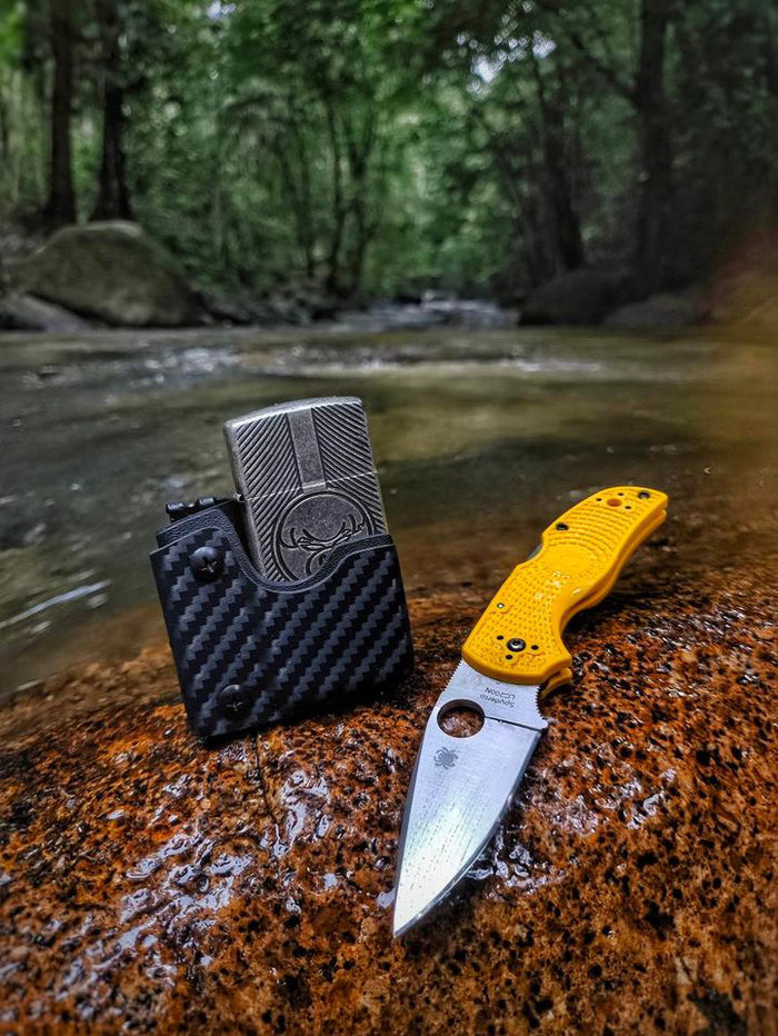 Outdoor lifestyle image of the carbon fiber TX2 KYDEX EDC Zippo lighter sheath beside a folding knife near a creek.