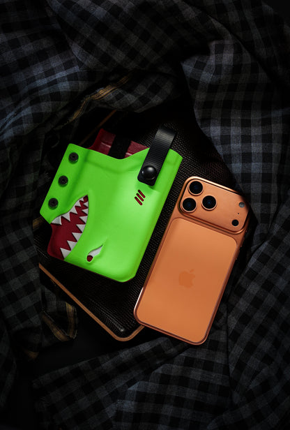 Neon green TACTICALGEEK TG EDC KYDEX shark phone sheath displayed beside an orange iPhone 17 Pro on a dark plaid background.