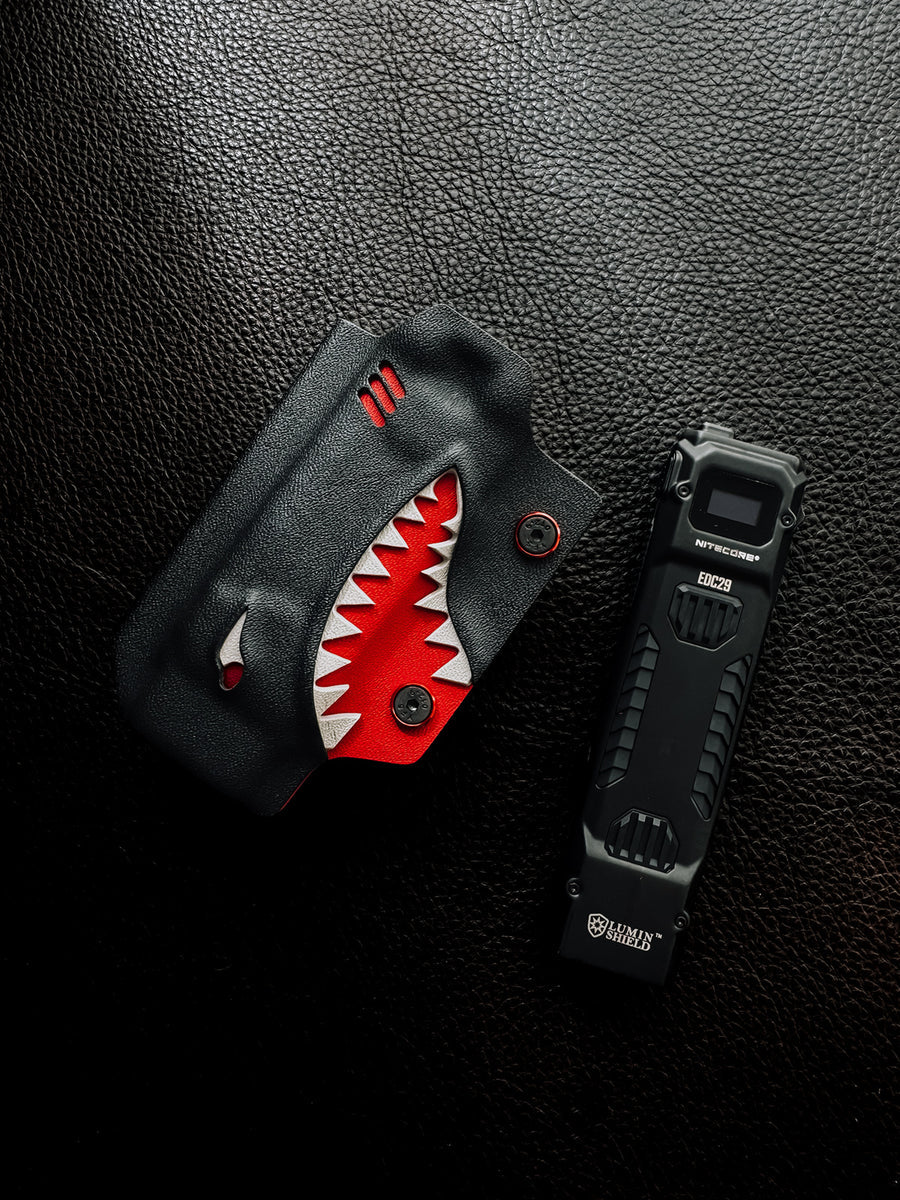Flat lay of the shark-style TX10 Flashlight Sheath placed beside a Nitecore EDC29 flashlight on a black leather surface.