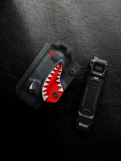 Flat lay of the shark-style TX10 Flashlight Sheath placed beside a Nitecore EDC29 flashlight on a black leather surface.