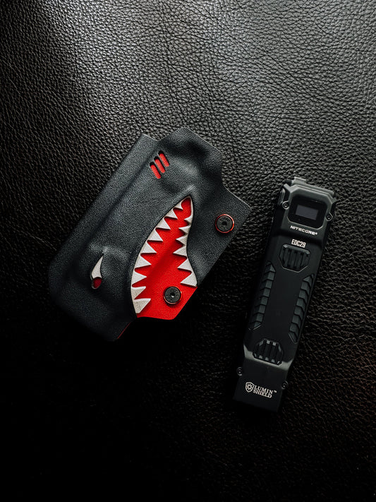 Flat lay of the shark-style TX10 Flashlight Sheath placed beside a Nitecore EDC29 flashlight on a black leather surface.