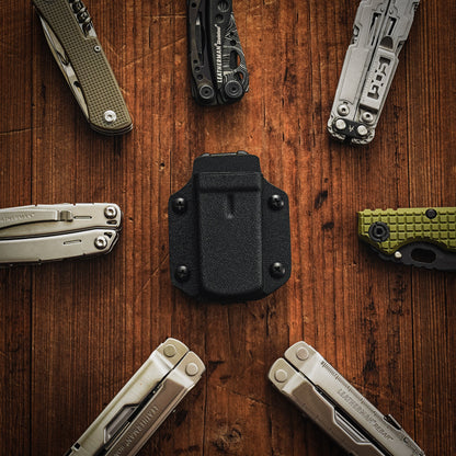 Flat lay image of the black TX3 Multitool Sheath centered on a wooden surface surrounded by multitools and folding knives.